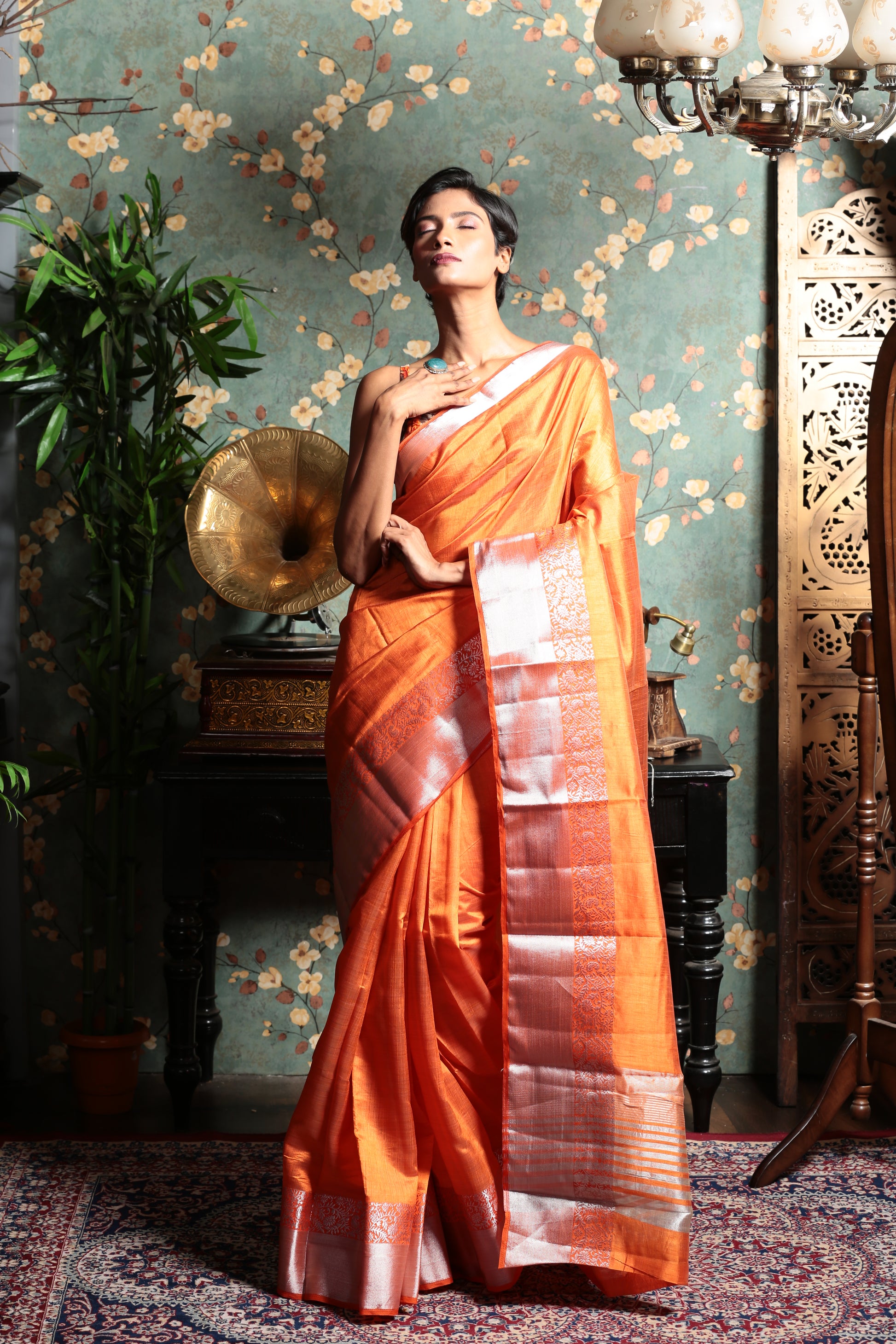 Model in a bright sunshine orange raw silk saree with a shiny silver patterned border, perfect for evenings | Urbane Essence