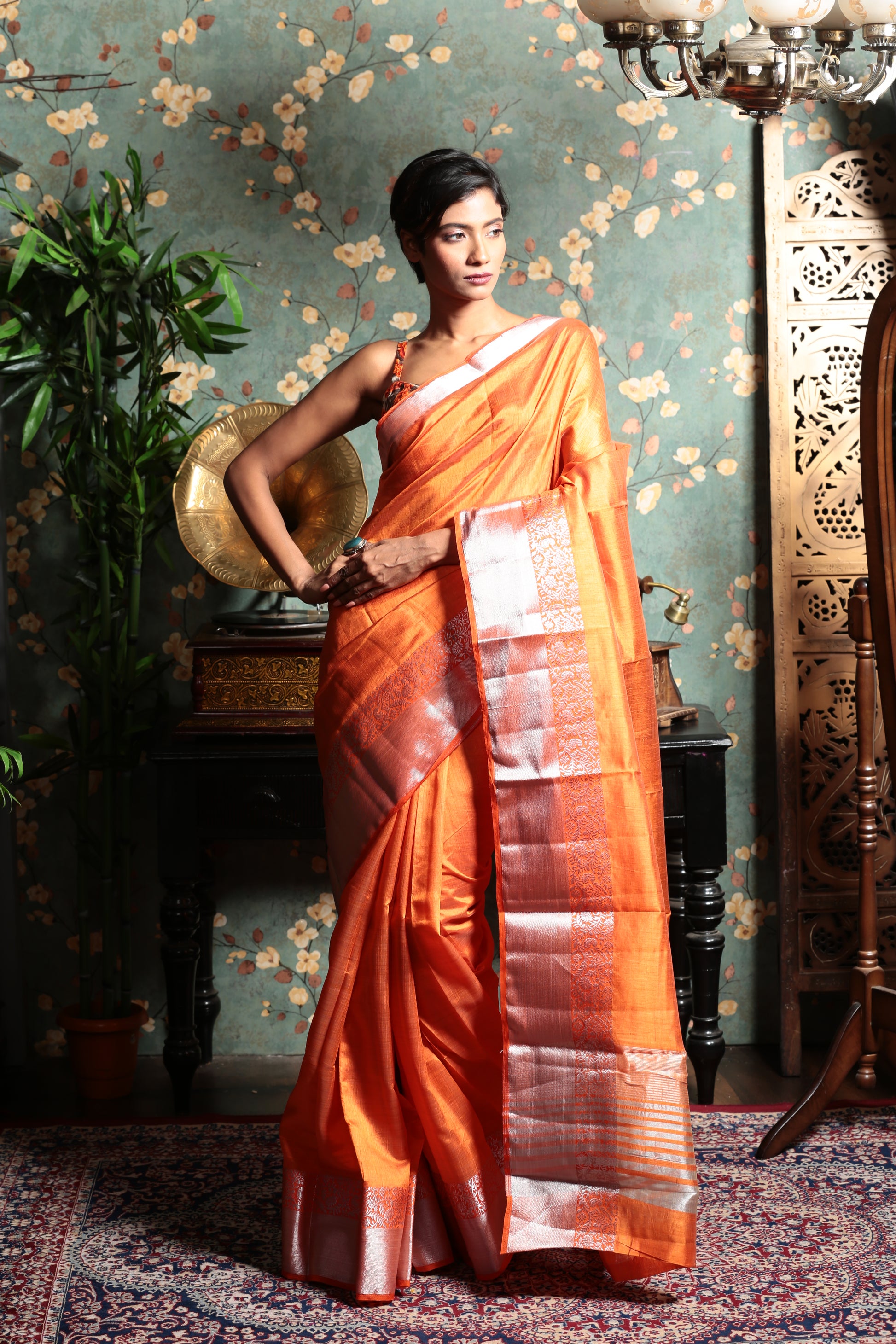 Bright orange raw silk saree with a shiny silver patterned border, perfect for evening wear | Urbane Essence